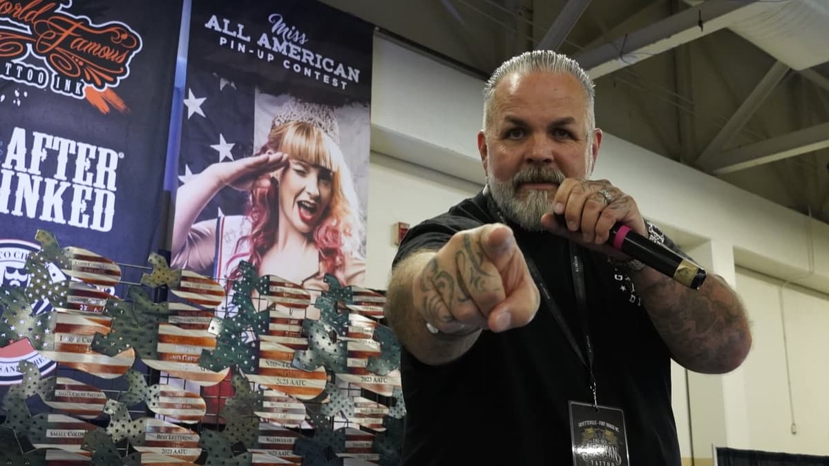 The host on the mic at the AATC show floor, pointing at the camera in front of the awards wall